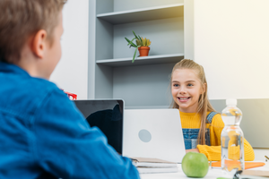 Junges Mädchen sitzt an einem Laptop und lacht einen Klassenkameraden an.