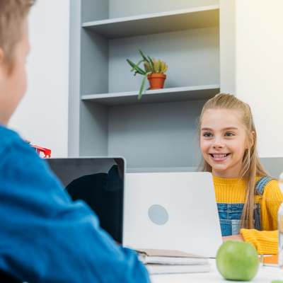 Junges Mädchen sitzt an einem Laptop und lacht einen Klassenkameraden an. 