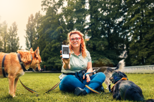 Frau sitzt auf einer Wiese und hat Hunde an der Leine. Sie h&auml;lt ein Smartphone mit der digitalen Hundemarke in die Kamera.